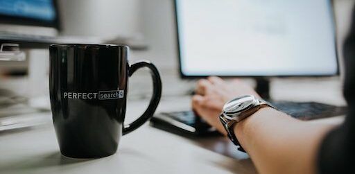 Coffee cup on desk next to hand on the keyboard of a computer
